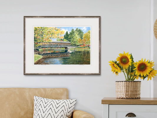 Framed watercolor print of Hemlock Bridge, Fryeburg, Maine, in a lifestyle setting with a beige sofa and sunflowers.