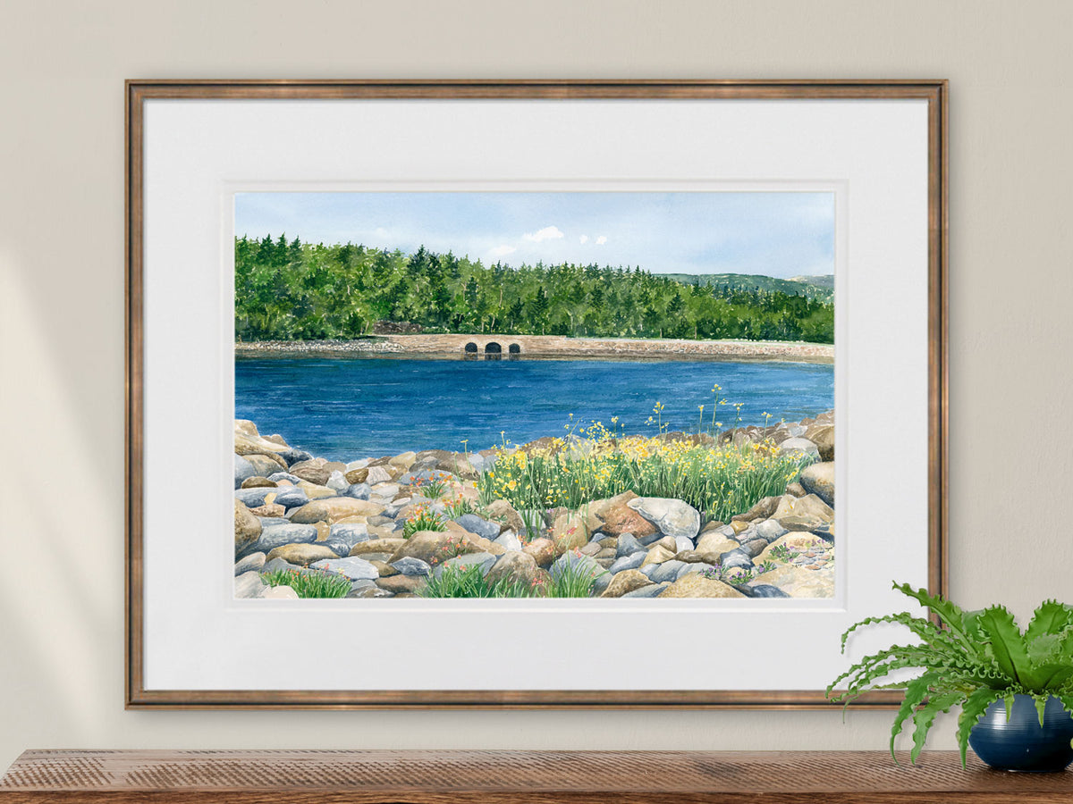 Framed original watercolor painting of Otter Cove Bridge, Acadia, in a warm gold frame over a wood table with a plant.