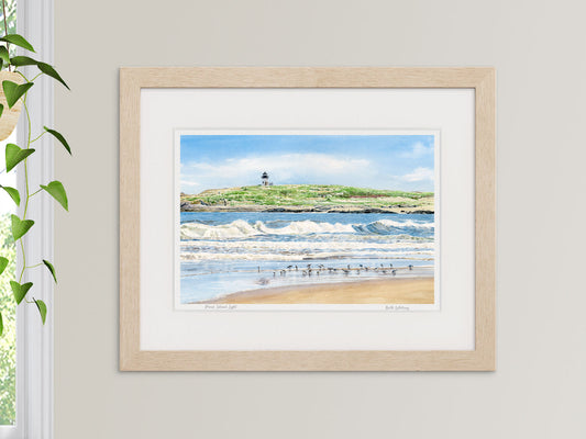 Framed watercolor print of Pond Island Light, Phippsburg, Maine, in a natural wood frame beside a window with a hanging plant.