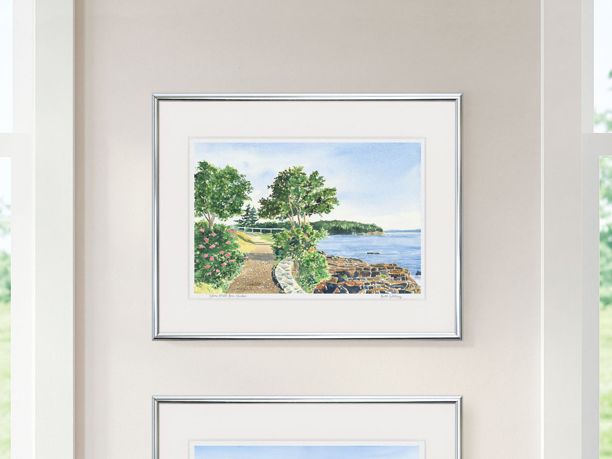Framed watercolor print of Shore Path Bar Harbor, Maine, in a silver frame as part of a diptych on a wall between windows.