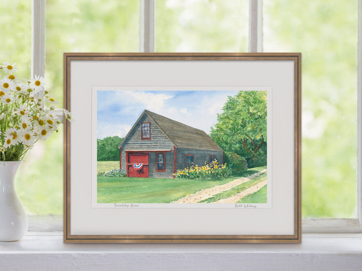 Framed watercolor print of Friendship Barn, Friendship, Maine, in a warm wood frame on a windowsill with a vase of flowers.