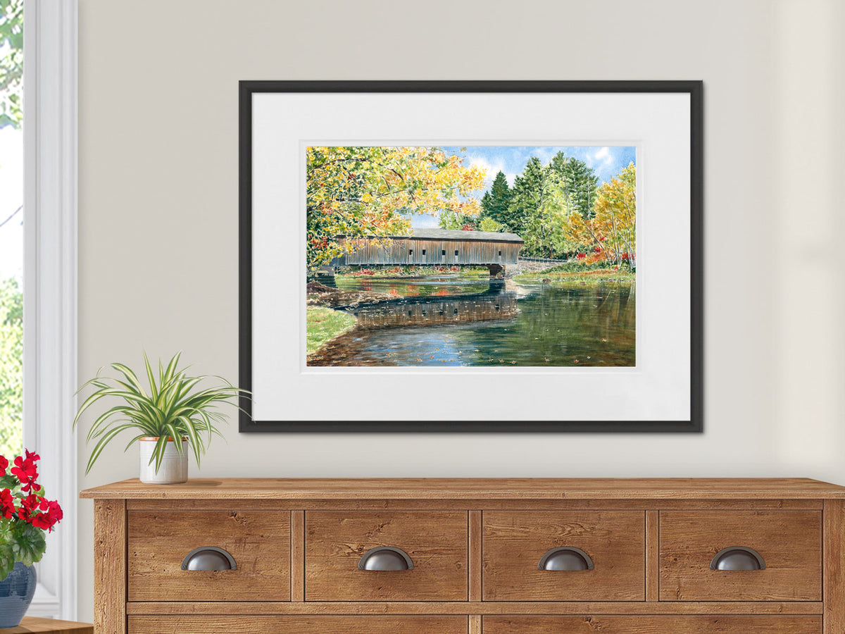 Framed painting of the Hemlock covered bridge in Fryeburg, Maine spanning a river with trees in the background, displayed in a black frame over a wooden sideboard.