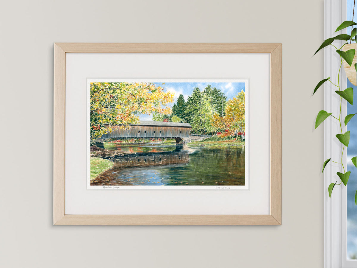 Framed watercolor print of Hemlock Bridge, Fryeburg, Maine beside a window with a hanging plant.