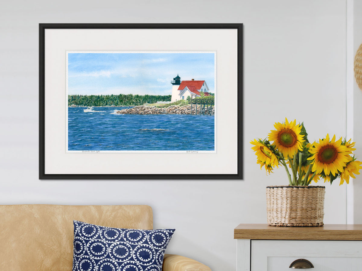 Framed watercolor print of Hendricks Head Light, Southport, Maine, in a black frame in a lifestyle setting with sunflowers.