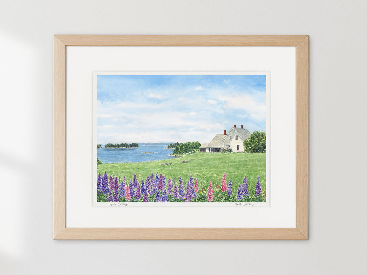 Framed watercolor print of Lupine Cottage, Deer Isle, Maine, in a warm wood frame on a neutral wall.