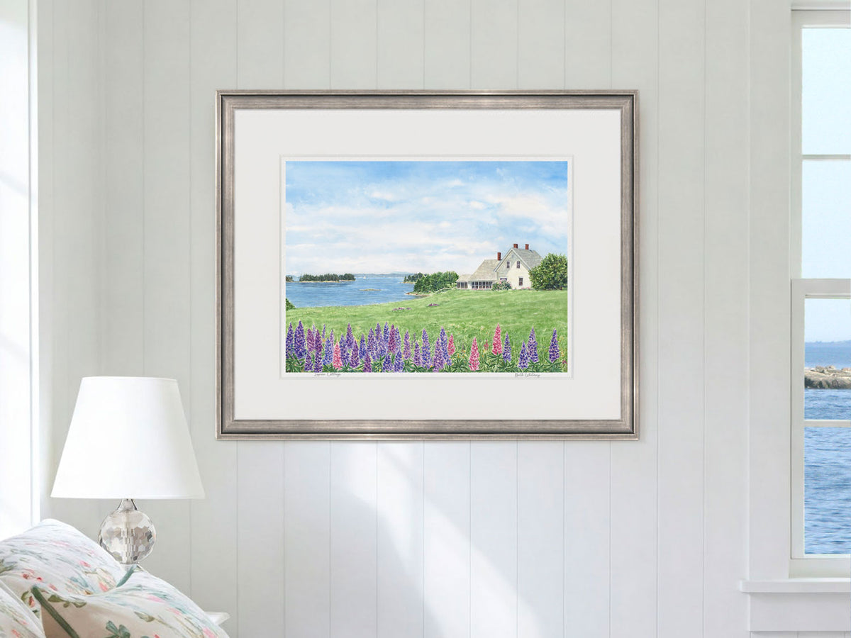 Framed watercolor print of Lupine Cottage, Deer Isle, Maine, in a silver frame in a bedroom lifestyle setting.