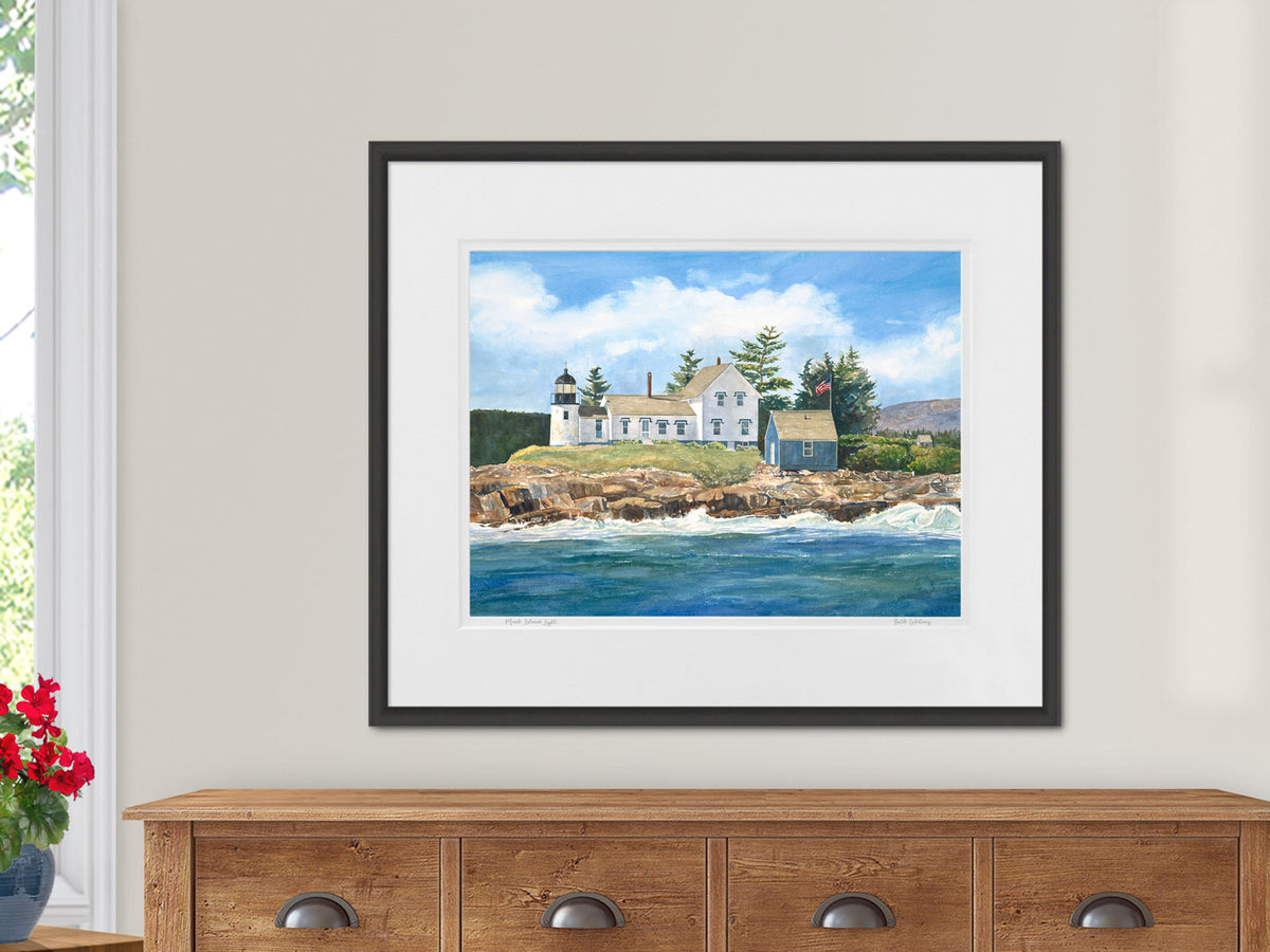 Framed watercolor print of Mark Island Light, Winter Harbor, Maine, in a black frame on a sideboard in a lifestyle setting.