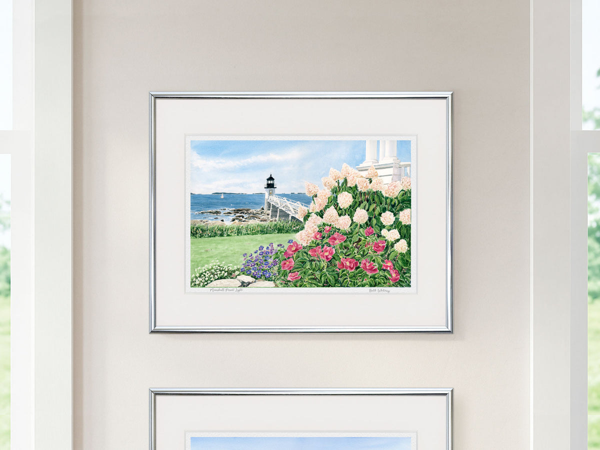 Framed watercolor print of Marshall Point Light in a silver frame on a neutral wall between windows, with another print below.