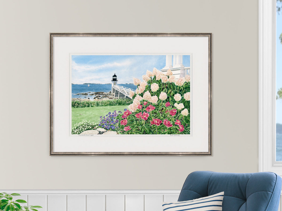 Framed watercolor print of Marshall Point Light in a gold frame in a nautical style sitting room with a window.