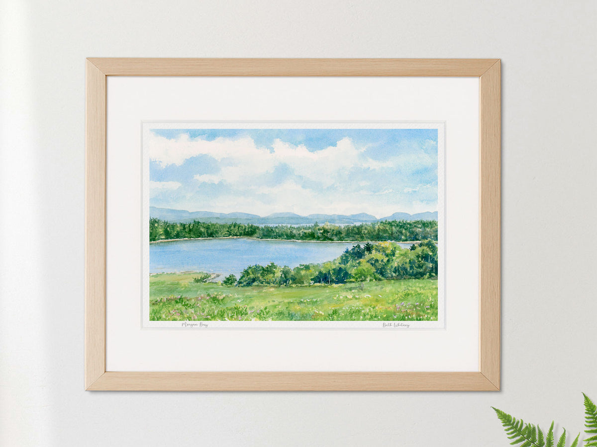 Framed watercolor print of Morgan Bay, Surry, Maine, in a warm wood frame on a neutral wall with a fern.