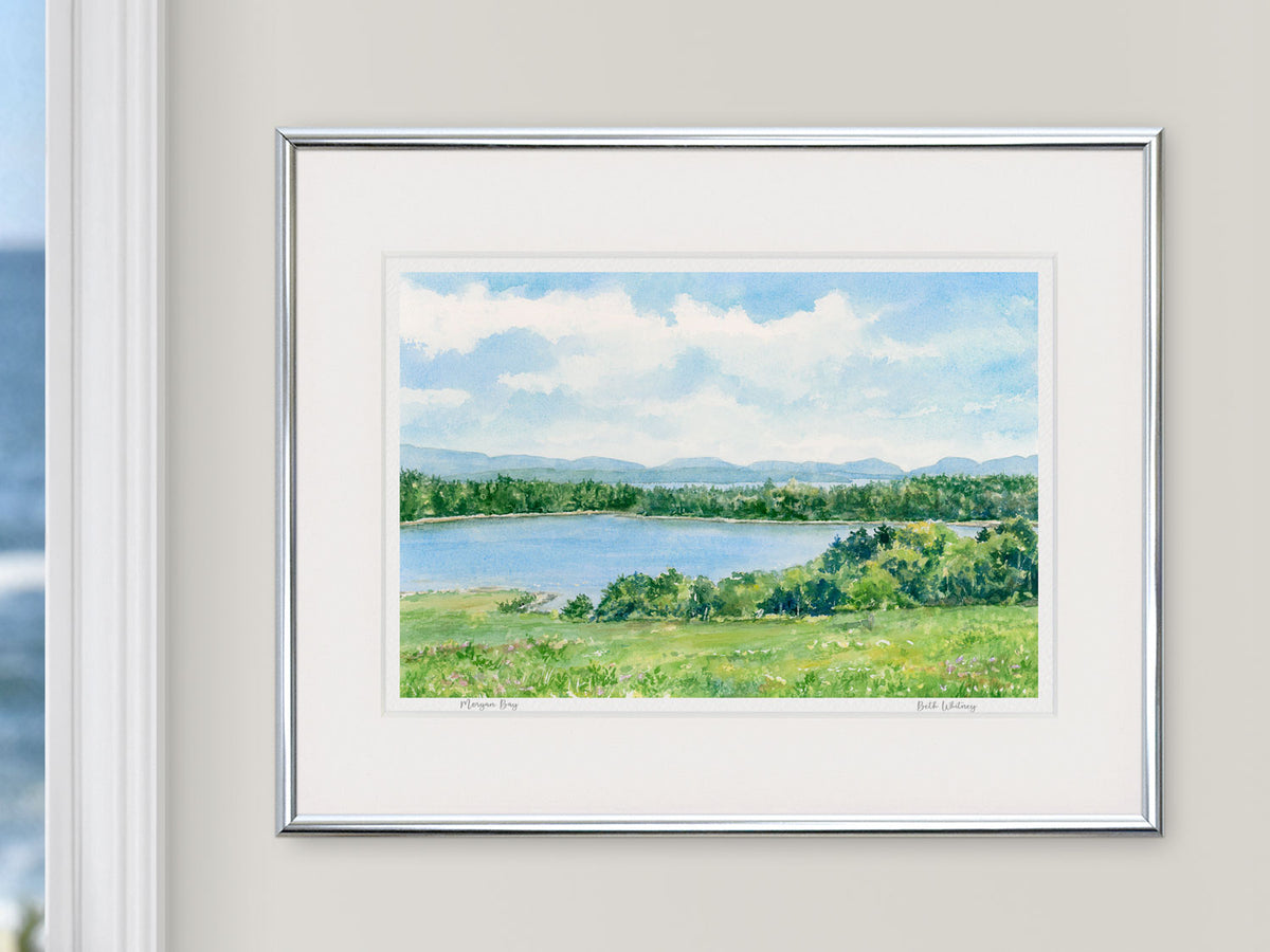 Framed watercolor print of Morgan Bay, Surry, Maine, in a simple silver frame beside a window with an ocean view.