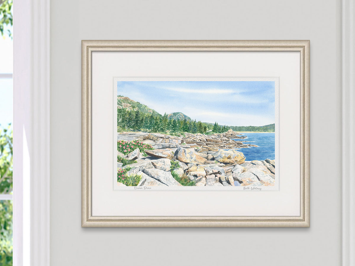 Framed watercolor print of Ocean Drive, Park Loop Road, Acadia, in a pale gold frame beside a sunny window.