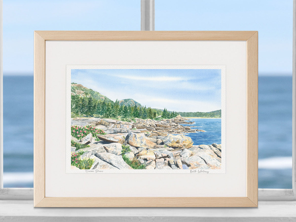 Framed watercolor print of Ocean Drive, Acadia National Park, in a natural wood frame in a coastal window setting.