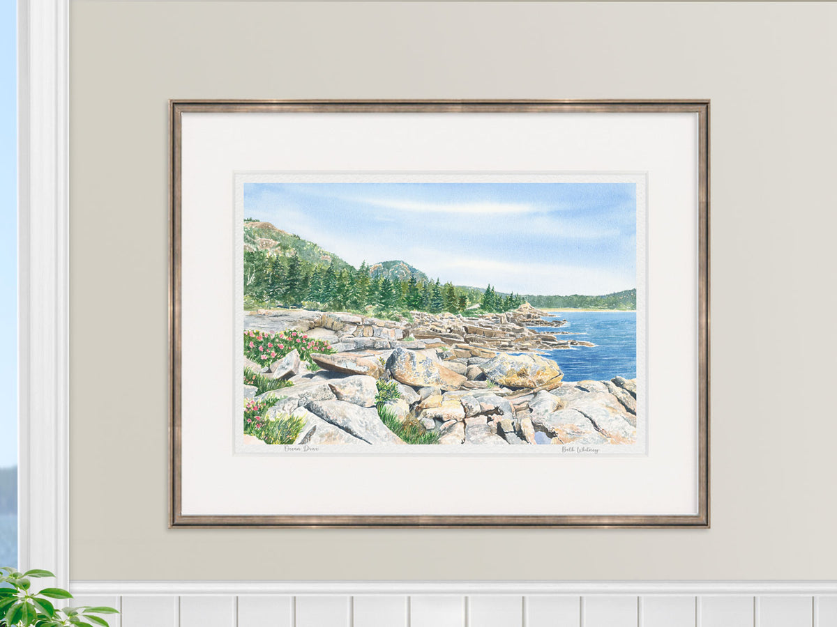 Framed watercolor print of Ocean Drive, Acadia, Maine, in a gold frame in a coastal home decor lifestyle setting.