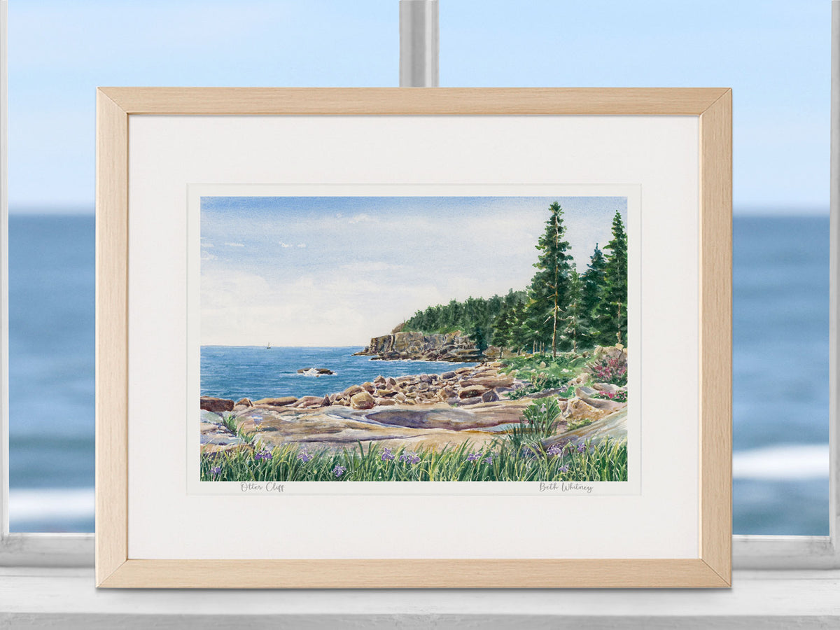 Framed watercolor print of Otter Cliff, Ocean Drive, Acadia, in a warm wood frame in a coastal decor window setting.
