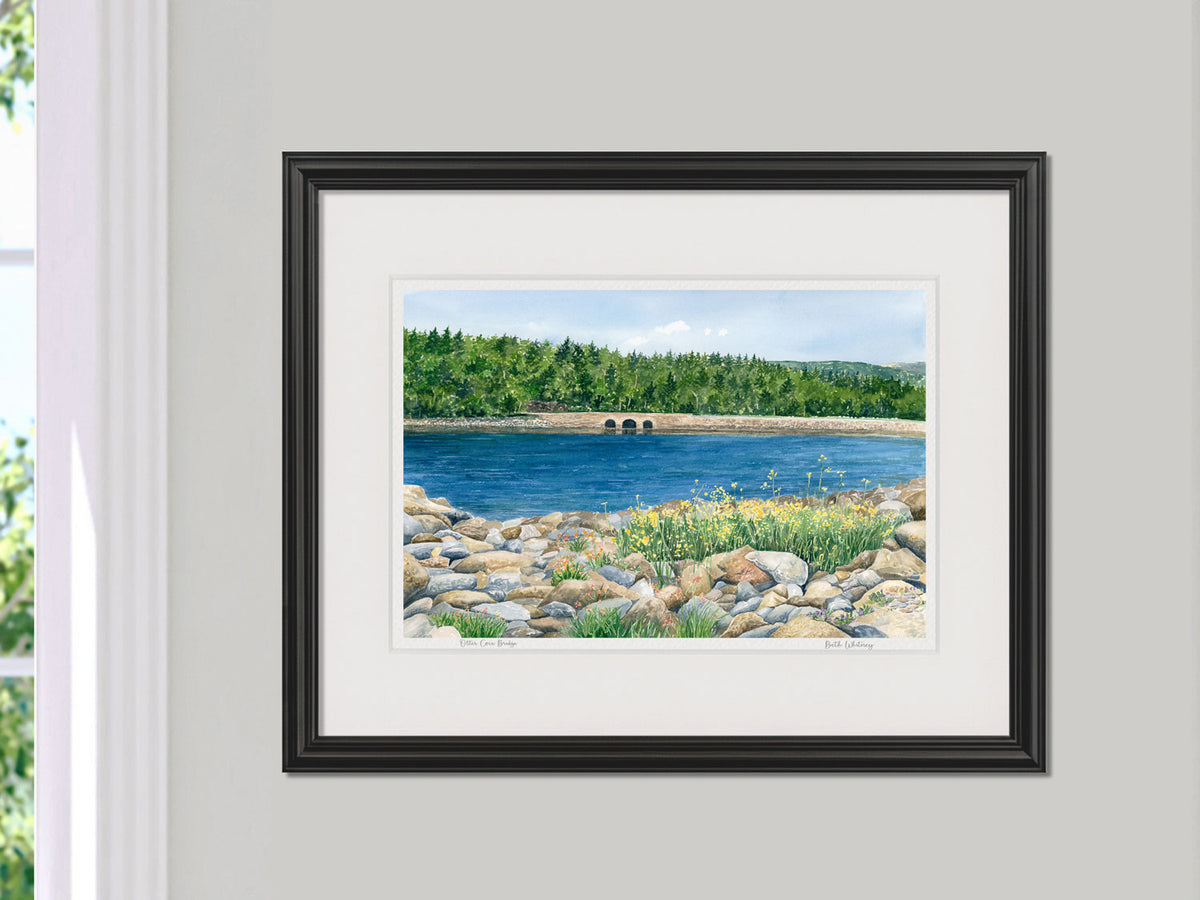Framed watercolor print of Otter Cove Bridge, Acadia, Maine, in a black frame beside a sunny window.