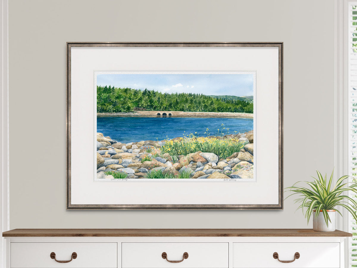Framed watercolor print of Otter Cove Bridge, Acadia, Maine, in a warm wood frame on a dresser in a lifestyle setting.