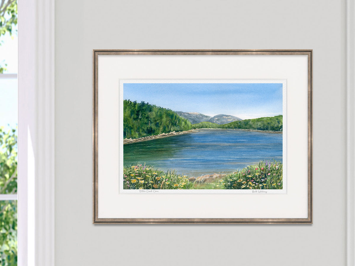 Framed watercolor print of Otter Creek Cove, Acadia, Maine, in a gold frame on a neutral wall beside a sunny window.