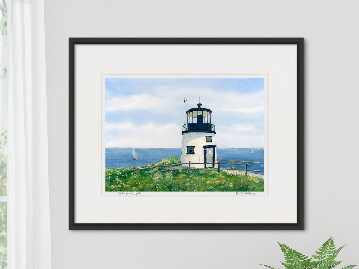 Framed watercolor print of Owls Head Light, Penobscot Bay, Maine, in a black frame on a neutral wall beside a window and fern.
