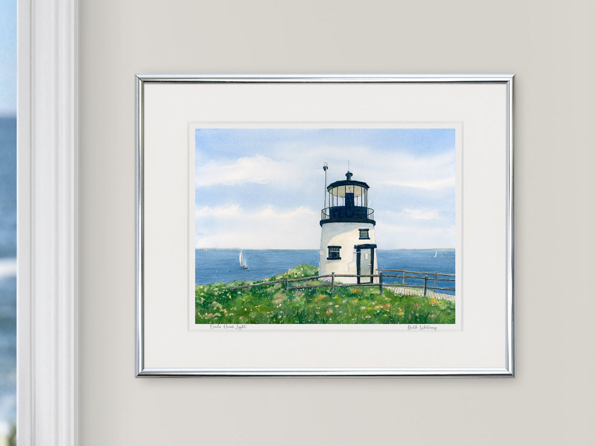Framed watercolor print of Owls Head Light, Midcoast Maine, in a silver frame on a neutral wall beside a window with an ocean view.
