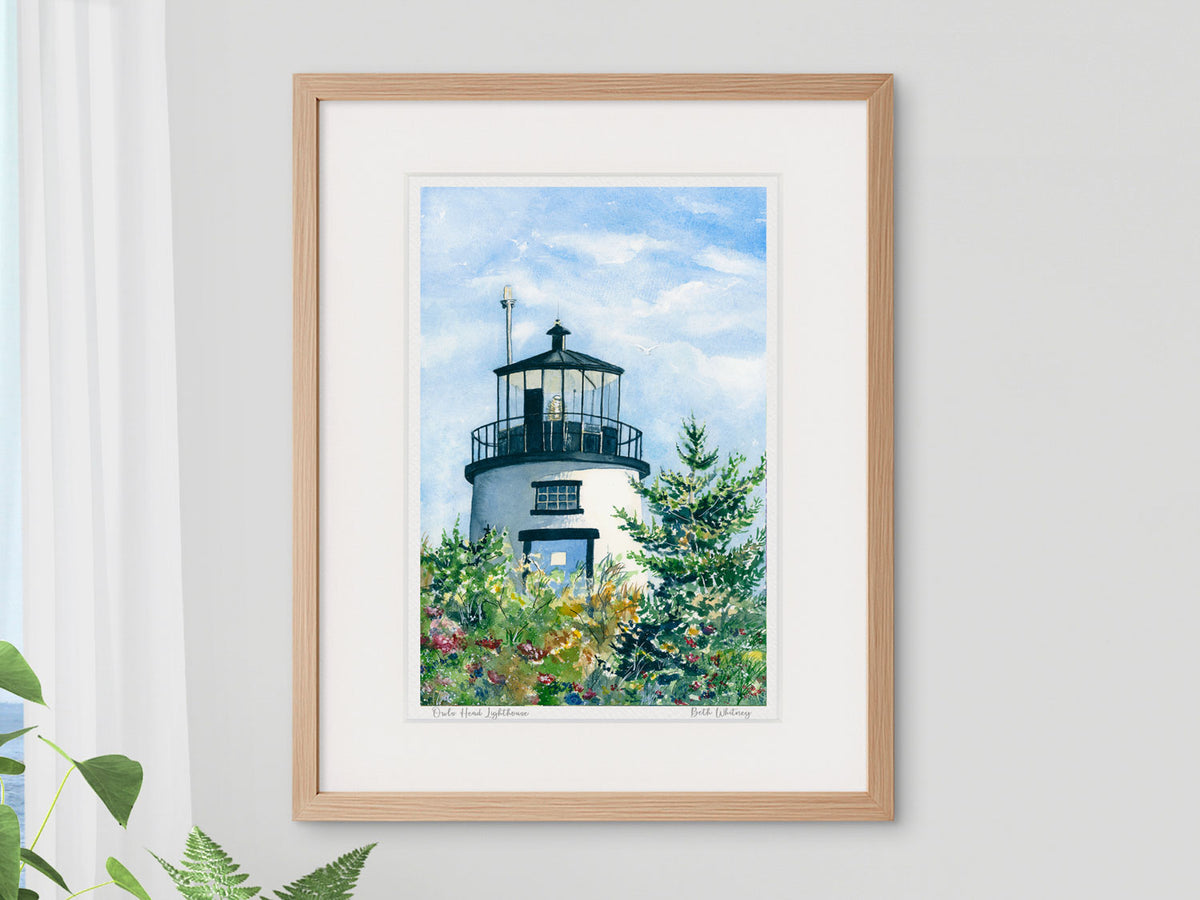 Framed watercolor print of Owls Head Lighthouse, Owls Head, Maine, in a warm wood frame with plant styling beside a window.