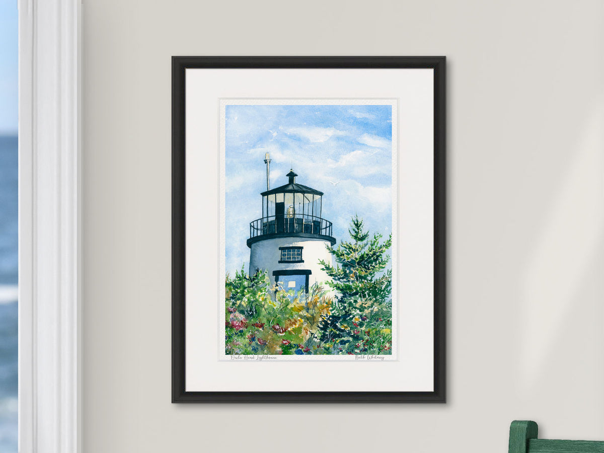 Framed watercolor print of Owls Head Lighthouse, Penobscot Bay, Maine, in a black frame beside a window with an ocean view and green chair.