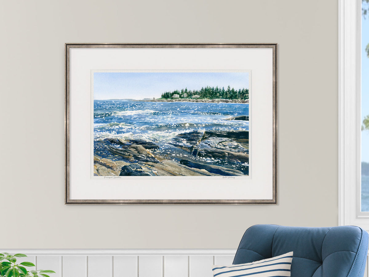 Framed watercolor print of Pemaquid Sparkles, Bristol, Maine, in a warm wood frame in a coastal bedroom lifestyle setting with a blue chair and striped pillow.
