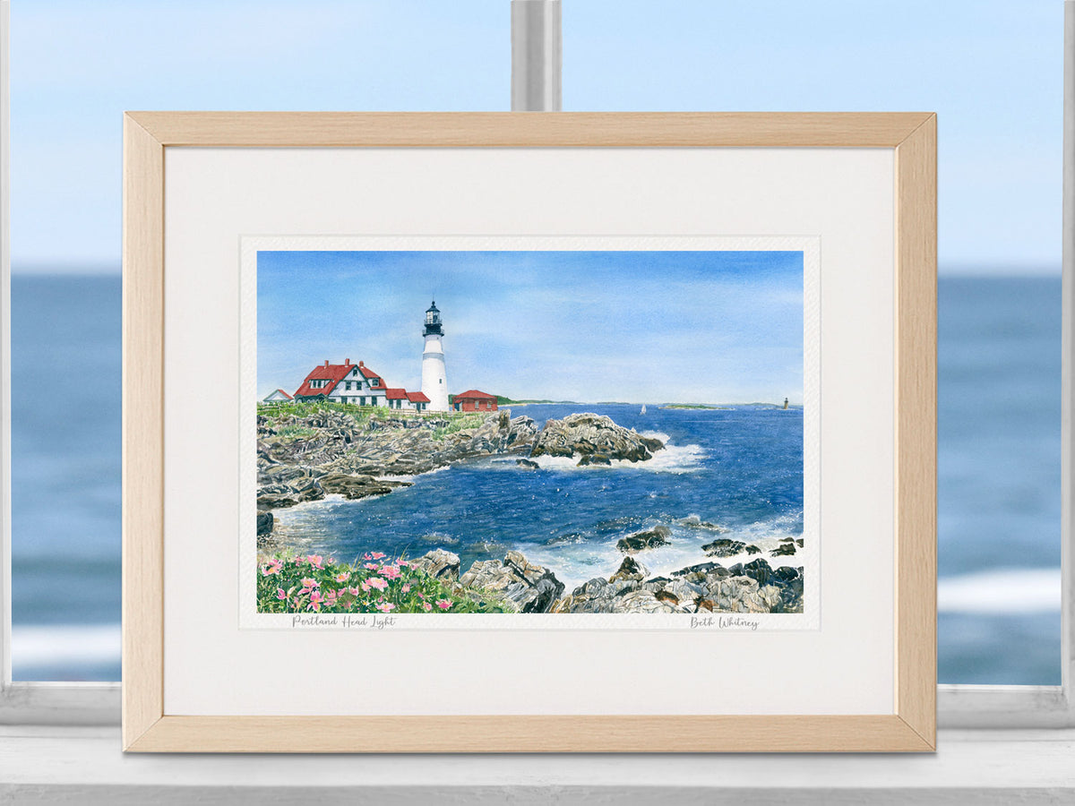 Framed watercolor print of Portland Head Light, Fort Williams Park, Maine, in a warm wood frame in a coastal window setting.