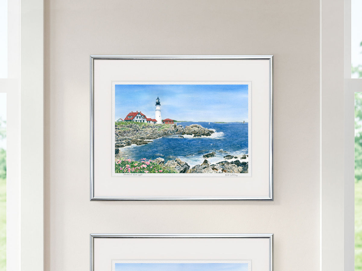 Framed watercolor print of Portland Head Light, Casco Bay, Maine, in a silver frame displayed between two windows on a neutral wall.