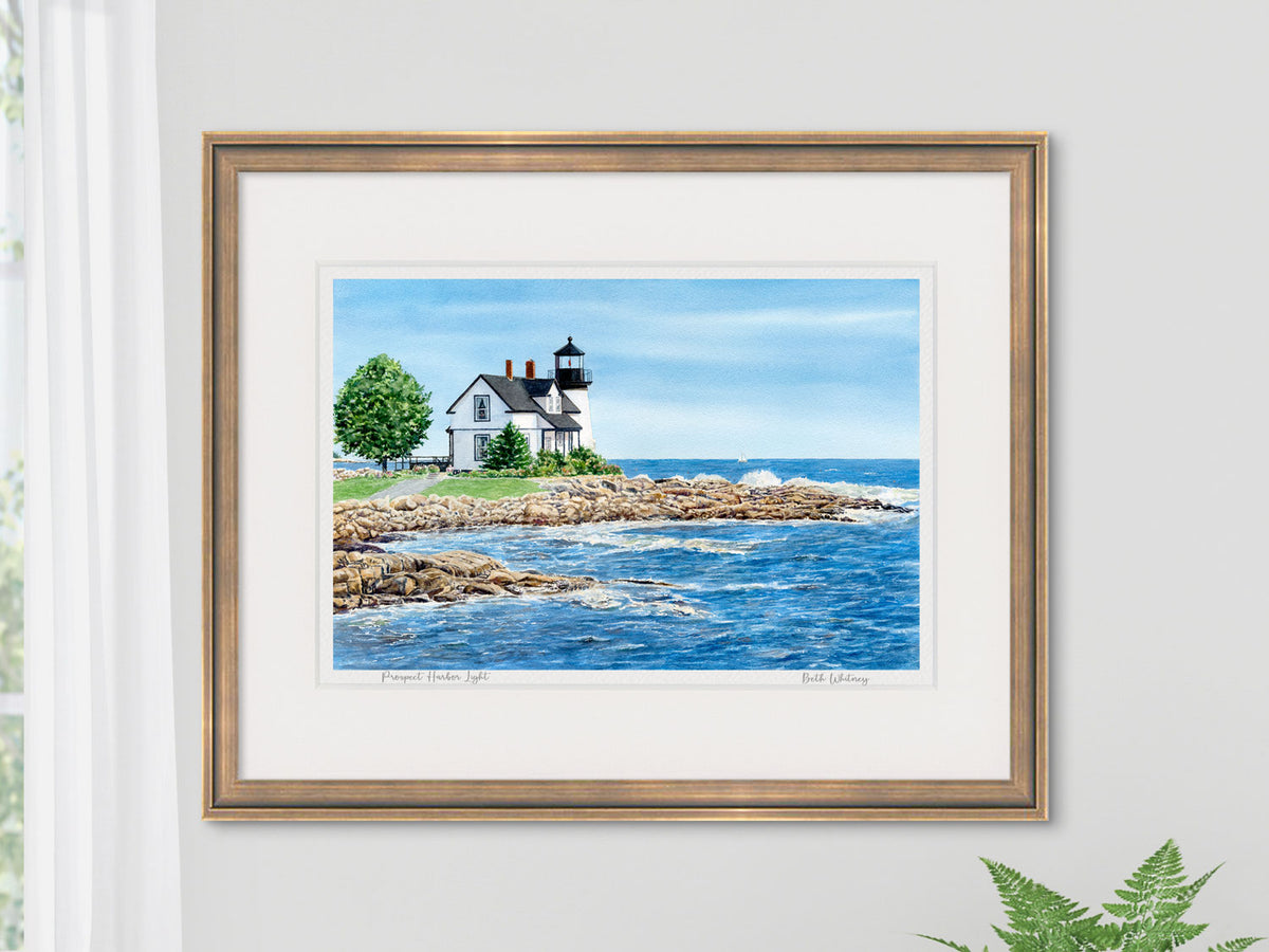 Framed watercolor print of Prospect Harbor Light, Bold Coast, Maine, in a gold frame with natural window light and fern houseplant.