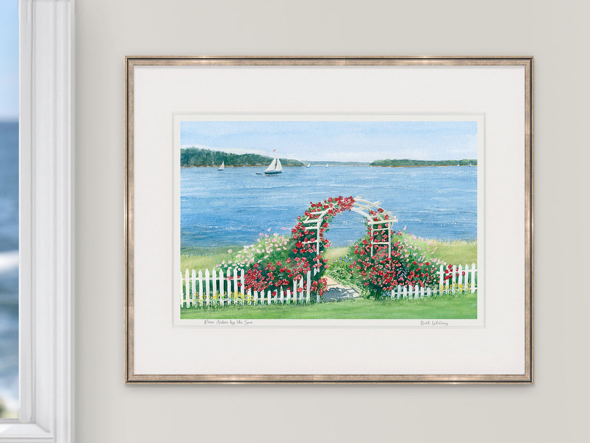 Framed watercolor print of Rose Arbor by the Sea, Brooksville, Maine, in a rose gold frame beside a window with an ocean view.