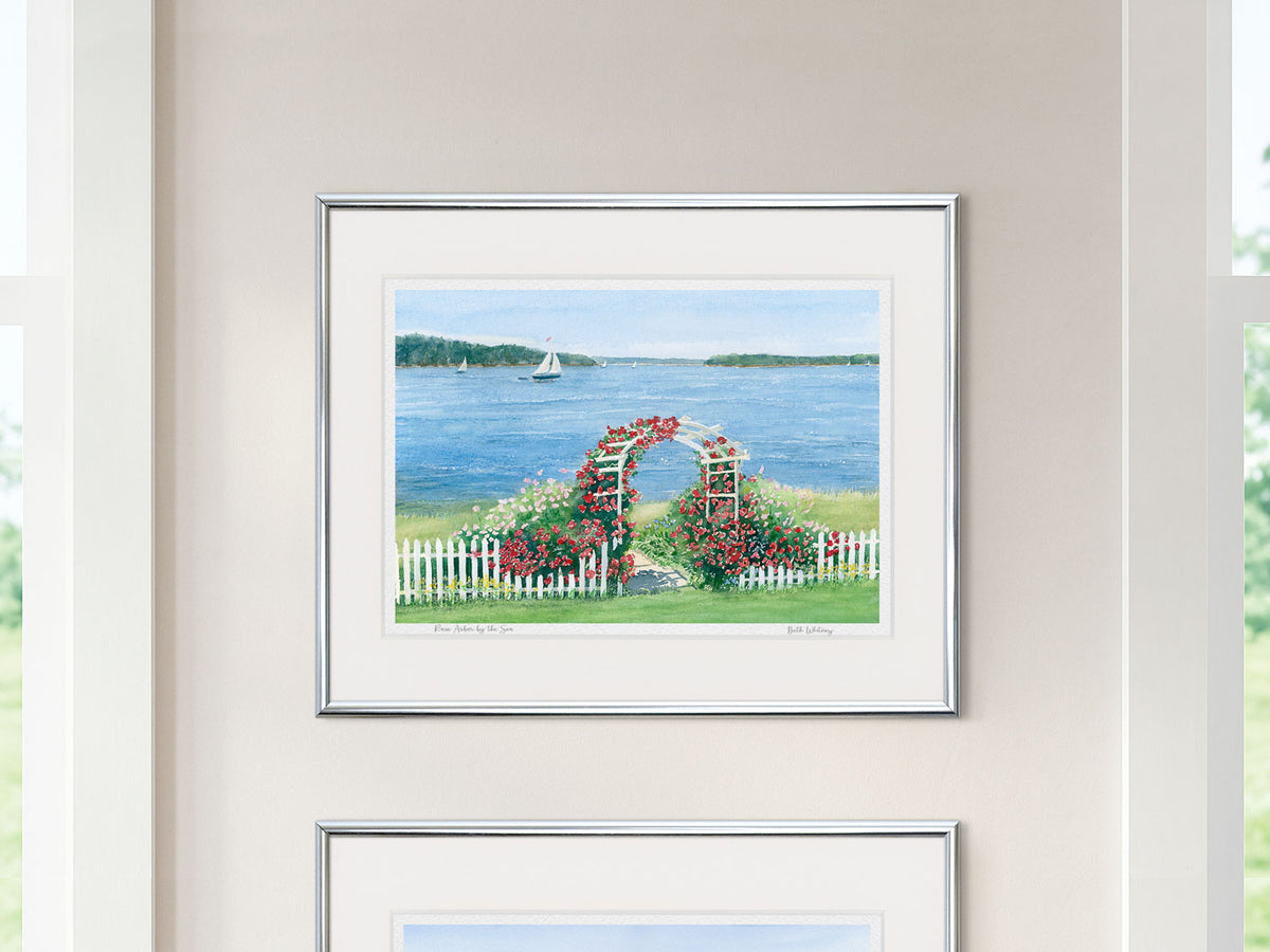 Framed watercolor print of Rose Arbor by the Sea, Penobscot Bay, in a silver frame on a neutral wall between two windows.