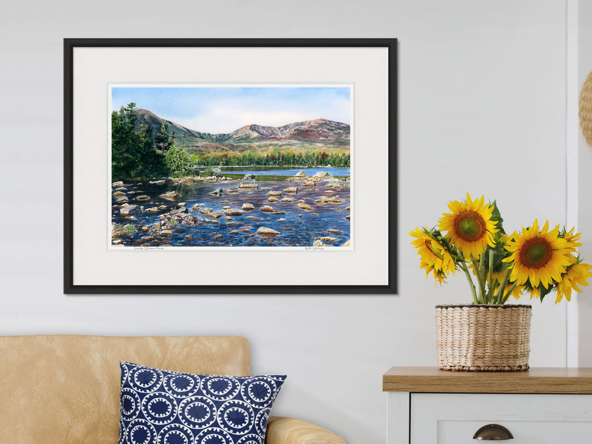 Framed watercolor print of Sandy Stream Pond, Mt Katahdin, Maine, in a black frame over a tan sofa with sunflower styling.