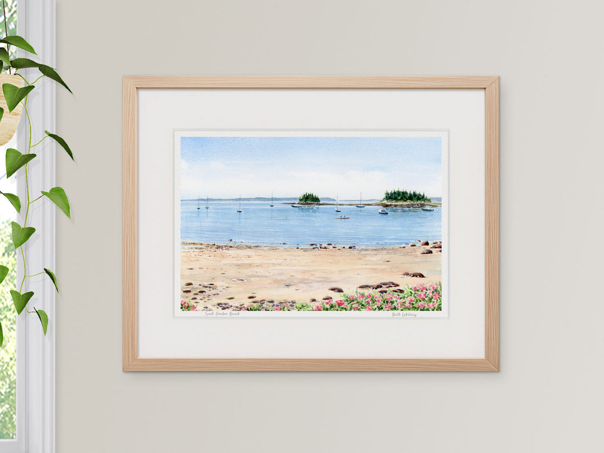 Framed watercolor print of Seal Harbor Beach, Seal Harbor, Maine, in a warm wood frame beside a window with a hanging plant.