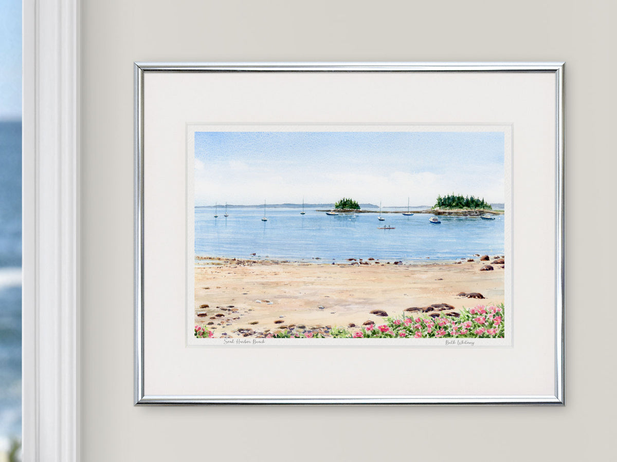 Framed watercolor print of Seal Harbor Beach, Acadia, Maine, in a silver frame beside a window with an ocean view.