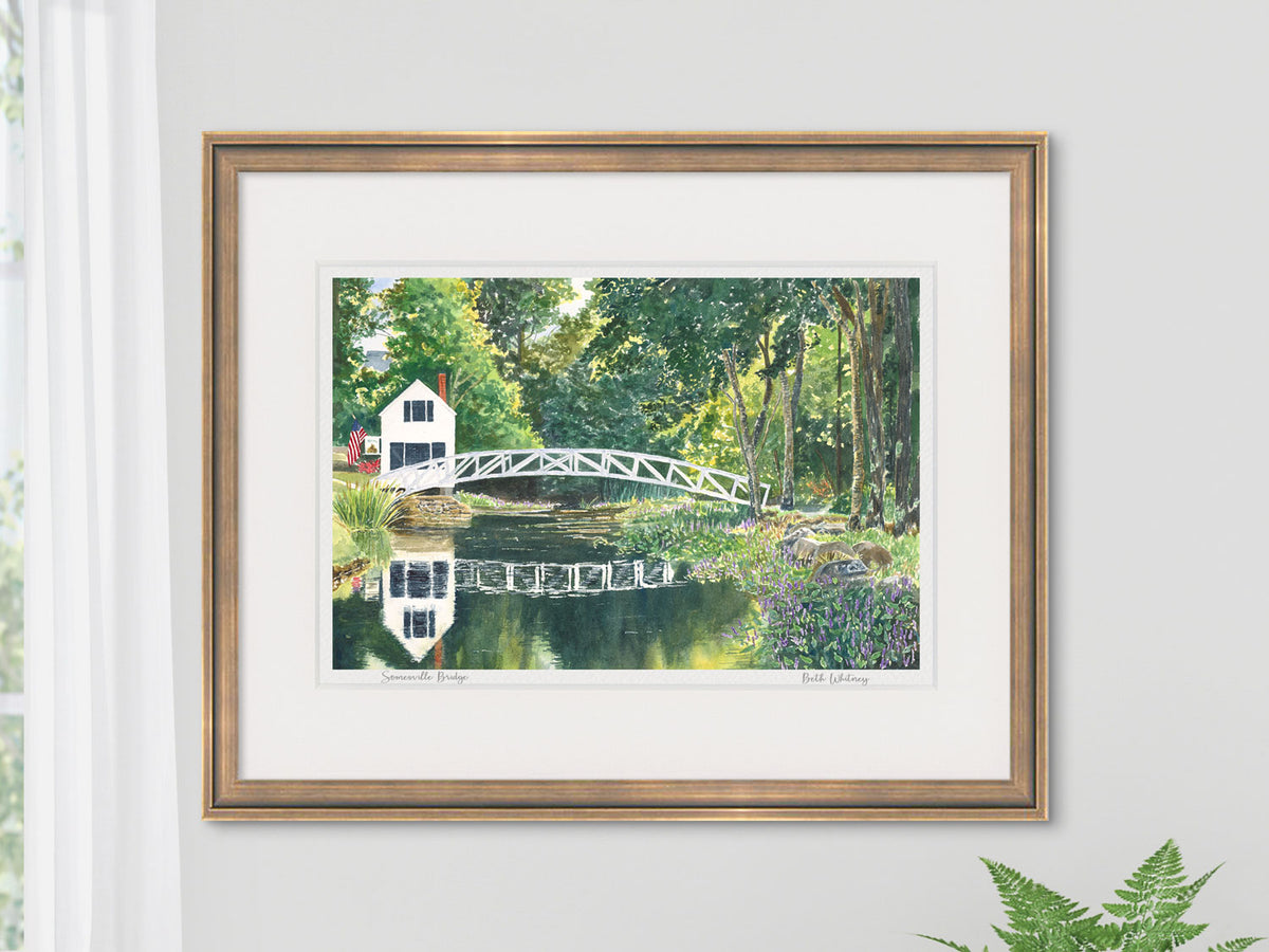 Framed watercolor print of Somesville Bridge, Somesville, Maine, in a gold frame on a cozy wall with a window and fern.