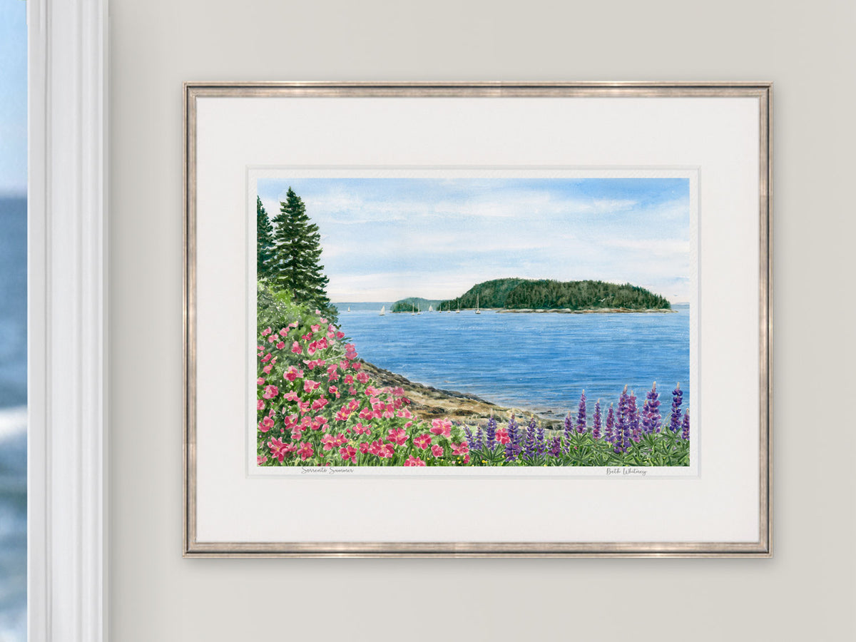 Framed watercolor print of Sorrento Summer in Downeast Maine, in a rose gold frame beside a window with an ocean view.