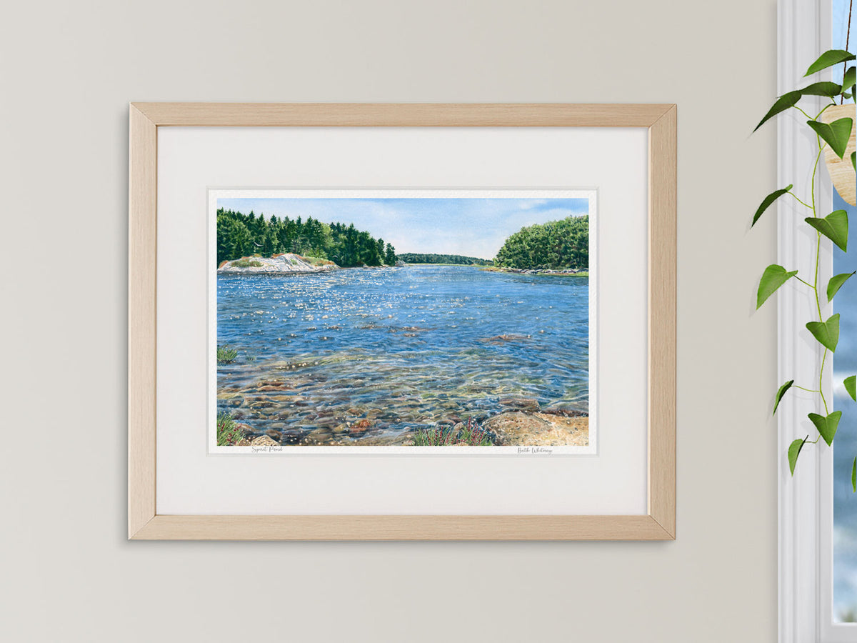 Framed watercolor print of Spirit Pond, Phippsburg, Maine, in a natural wood frame beside a window with a hanging plant and ocean view.