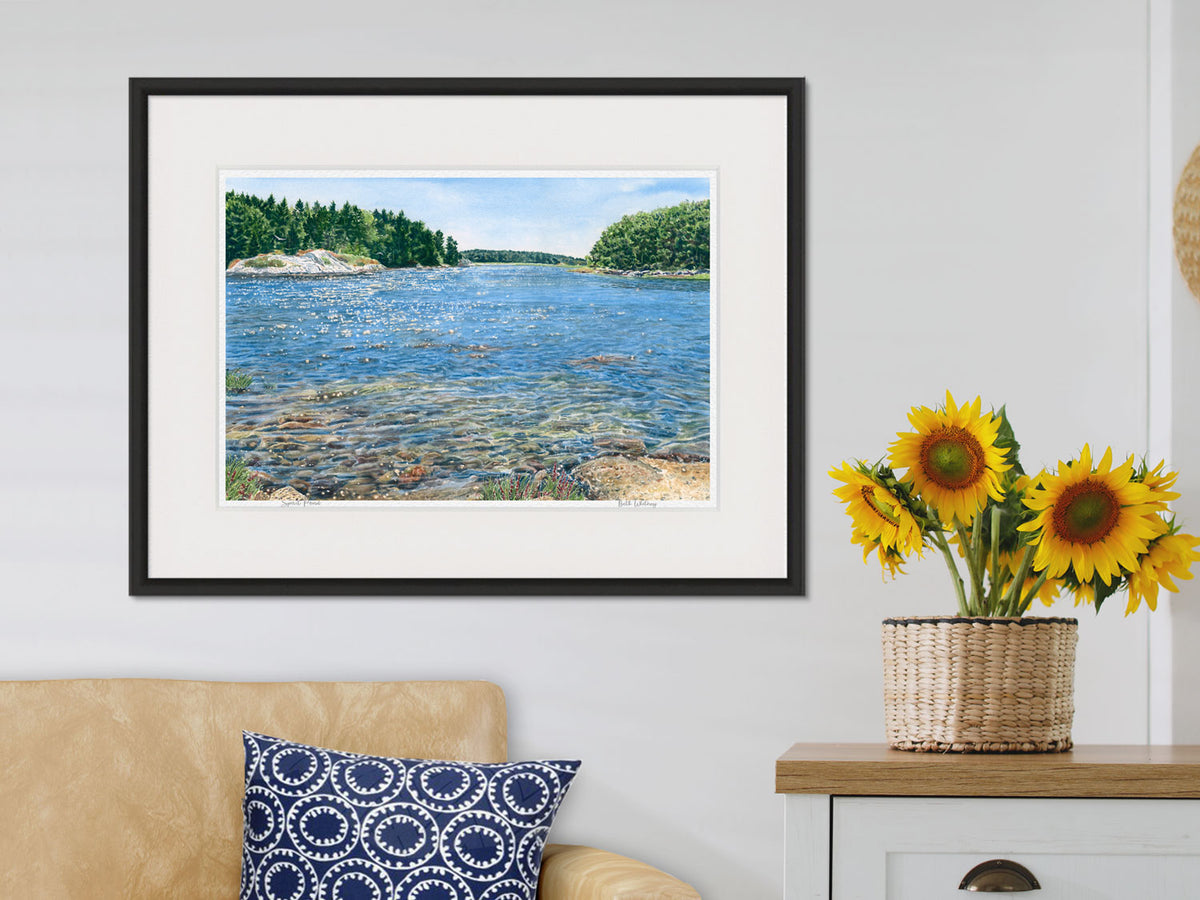 Framed watercolor print of Spirit Pond, Phippsburg, Maine, in a striking black frame in a cozy living room with sunflowers.