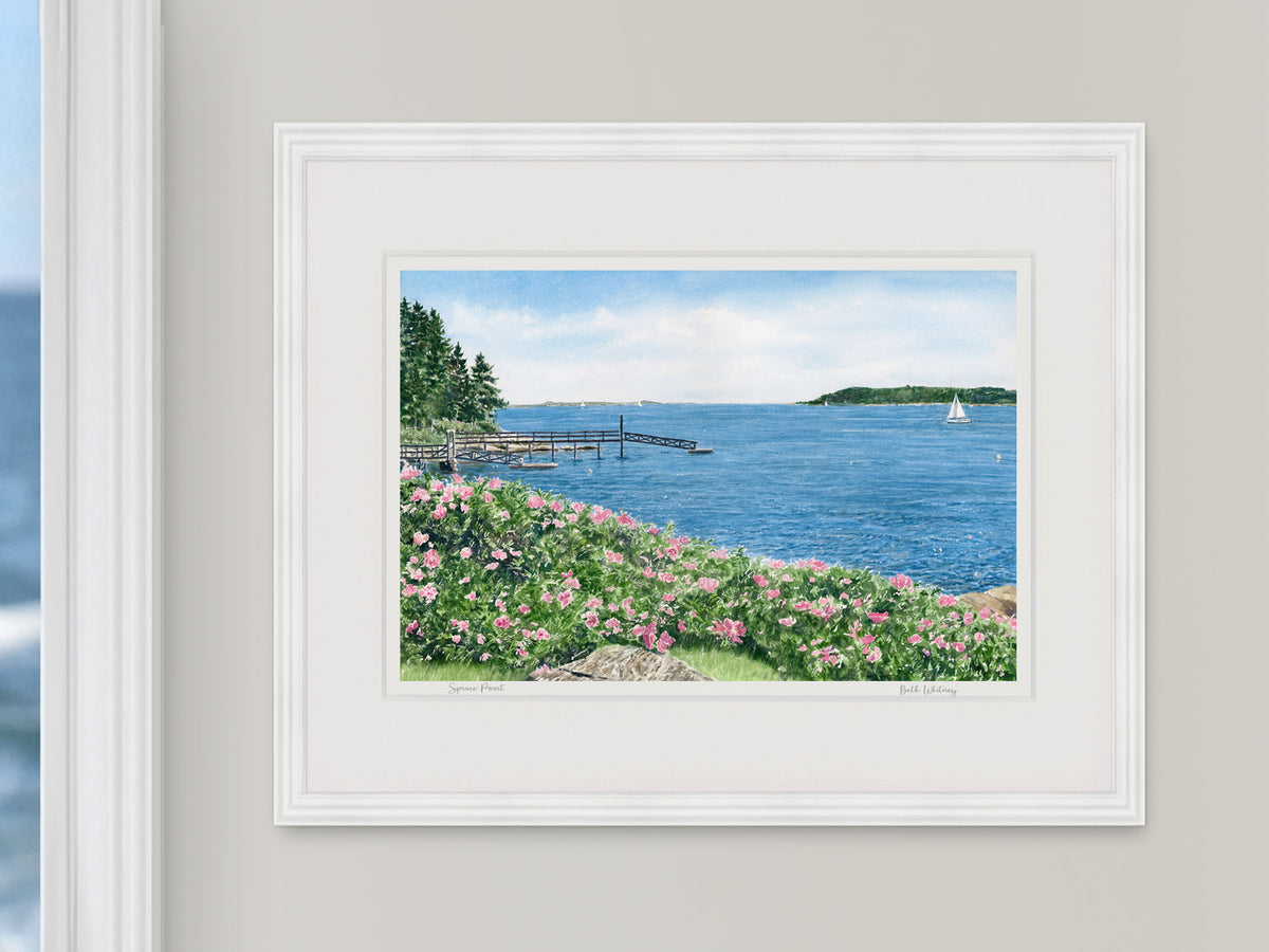 Framed watercolor print of Spruce Point, Boothbay Harbor, in a white frame on a light wall beside a window with an ocean view.
