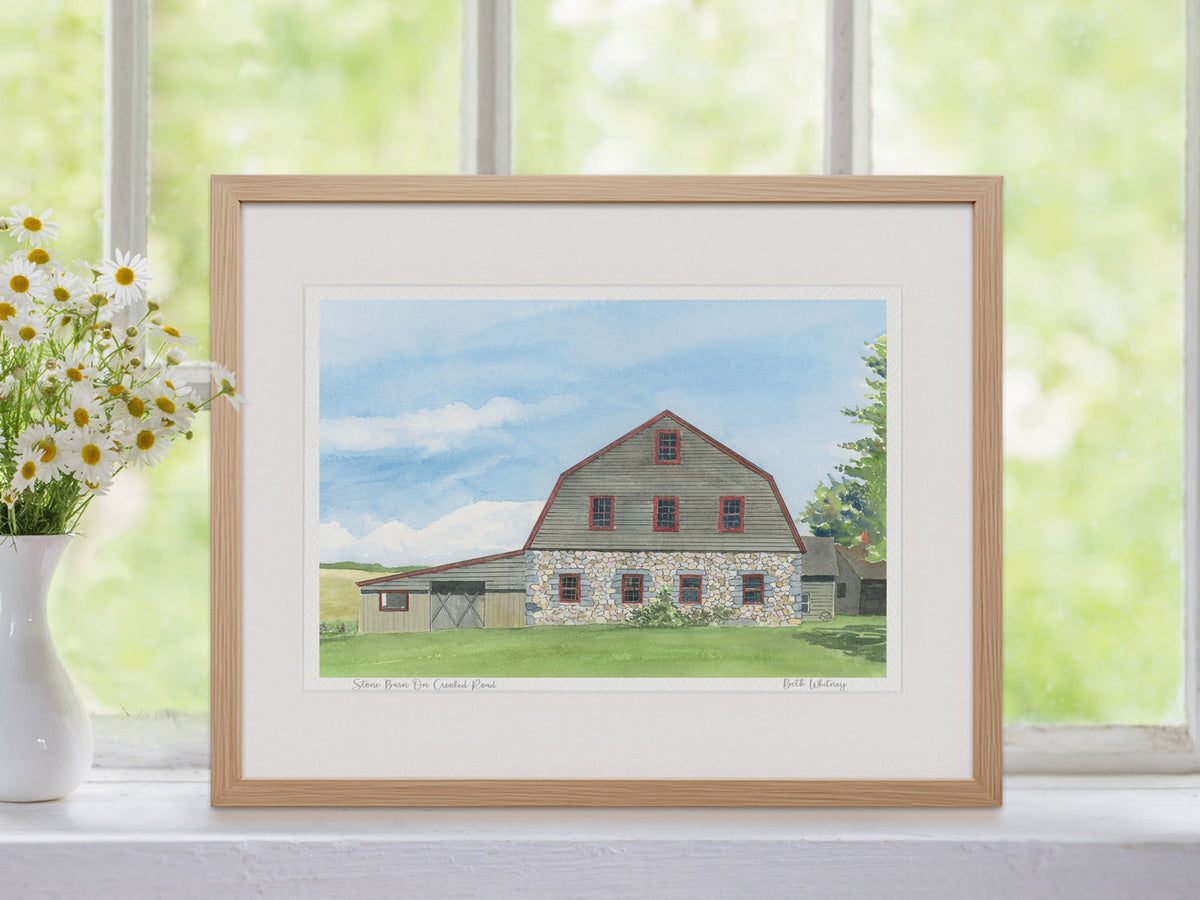 Framed watercolor print of Stone Barn on Crooked Road, Mount Desert Island, Maine, in a warm wood frame in a summer window setting.
