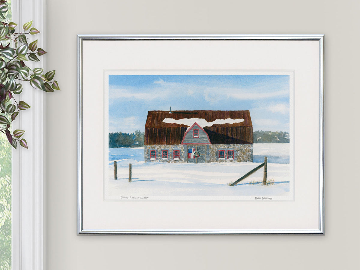Framed watercolor print of Stone Barn in Winter, Bar Harbor, Maine, in a silver frame beside a window with a hanging plant.
