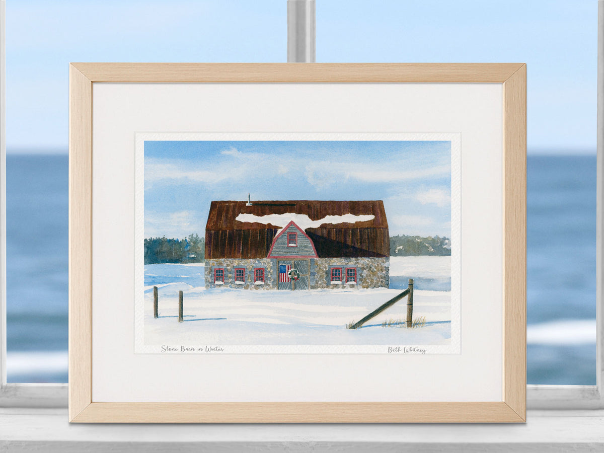 Framed watercolor print of Stone Barn in Winter, Bar Harbor, Maine, in a natural wood frame on a in a coastal windowsill setting.