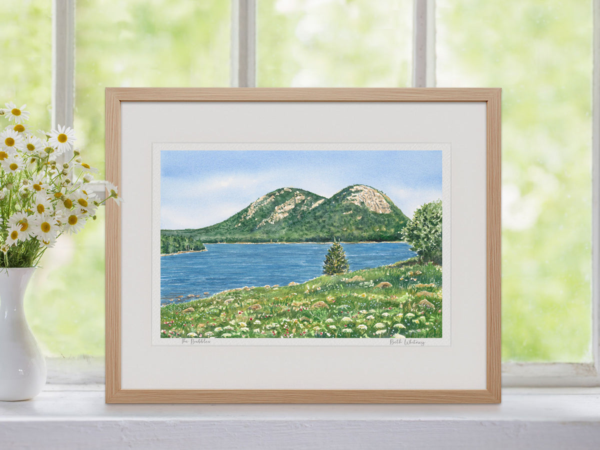 Framed watercolor print of The Bubbles, Jordan Pond, Acadia, in a warm wood frame in a window setting with wildflowers.