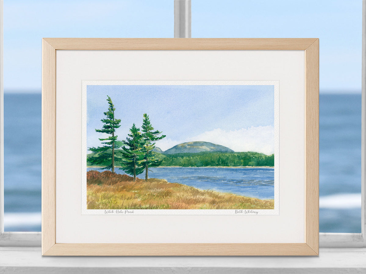 Framed watercolor print of Witch Hole Pond, Bar Harbor, Maine, in a warm wood frame in a coastal window setting.