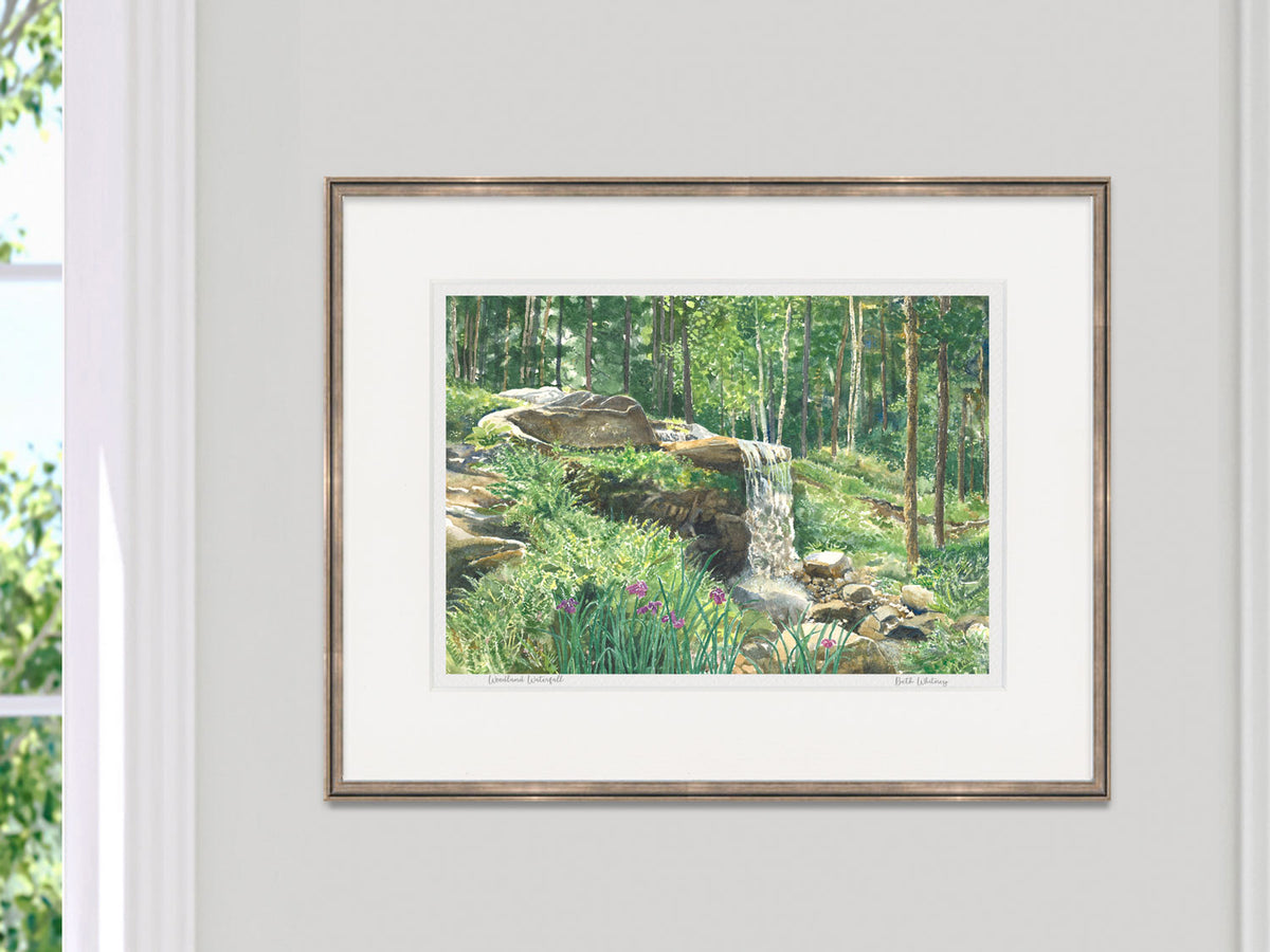 Framed watercolor print of Woodland Waterfall, Boothbay, Maine, in a gold frame beside a sunny window.