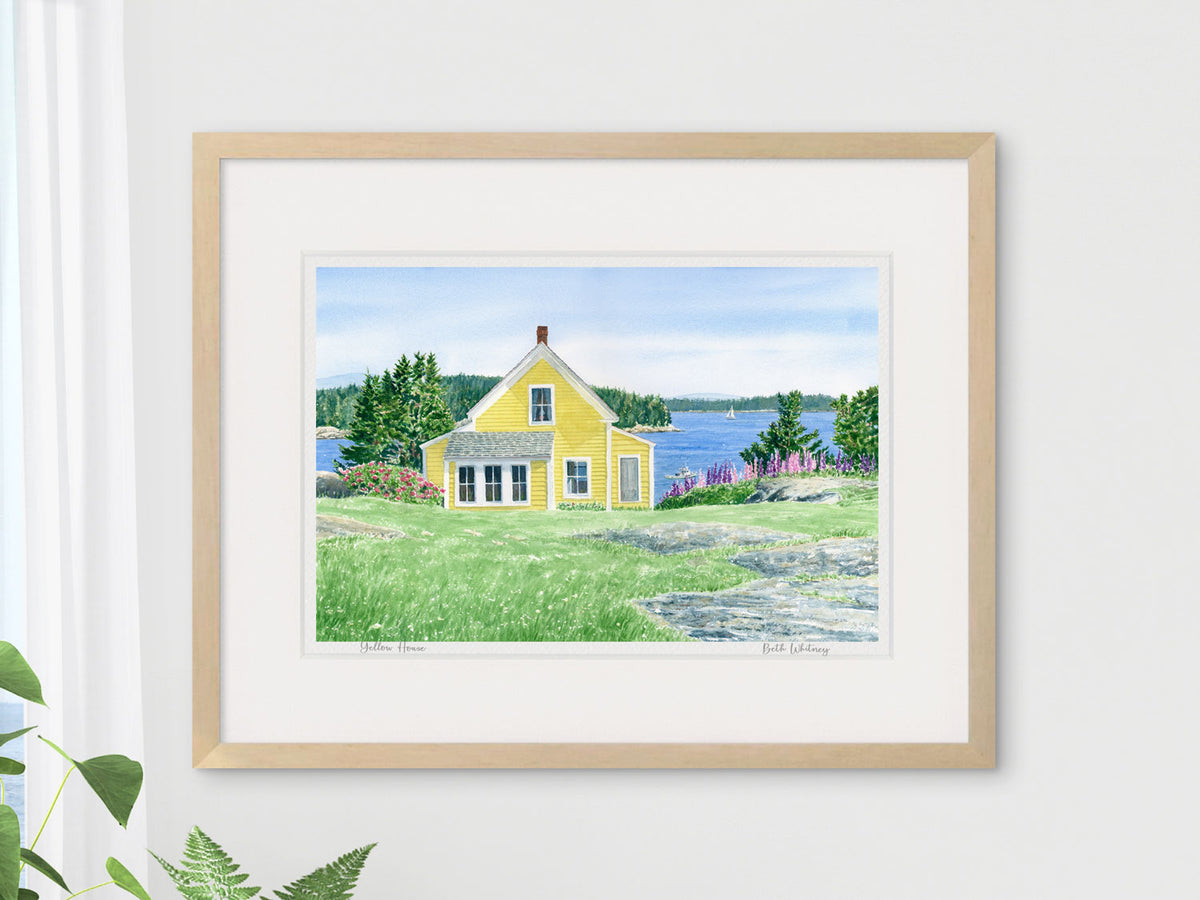 Framed watercolor print of Yellow House, Deer Isle, Maine, in a wood frame beside a window with houseplant styling.