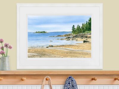 Watercolor painting of sandy beach cove and ocean, shown in white frame over a coatrack