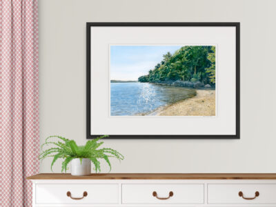 Watercolor print of sparkly ocean cove with beach, rocky shoreline, and trees shown over a white dresser