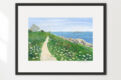 Watercolor of seaside cottage with path along the ocean, winding through grass and wildflowers, shown in a black frame.
